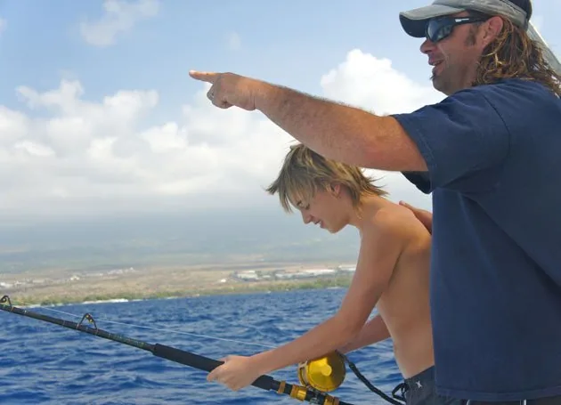 Captain pointing along the Kona coastline while a young angler holds a fishing rod